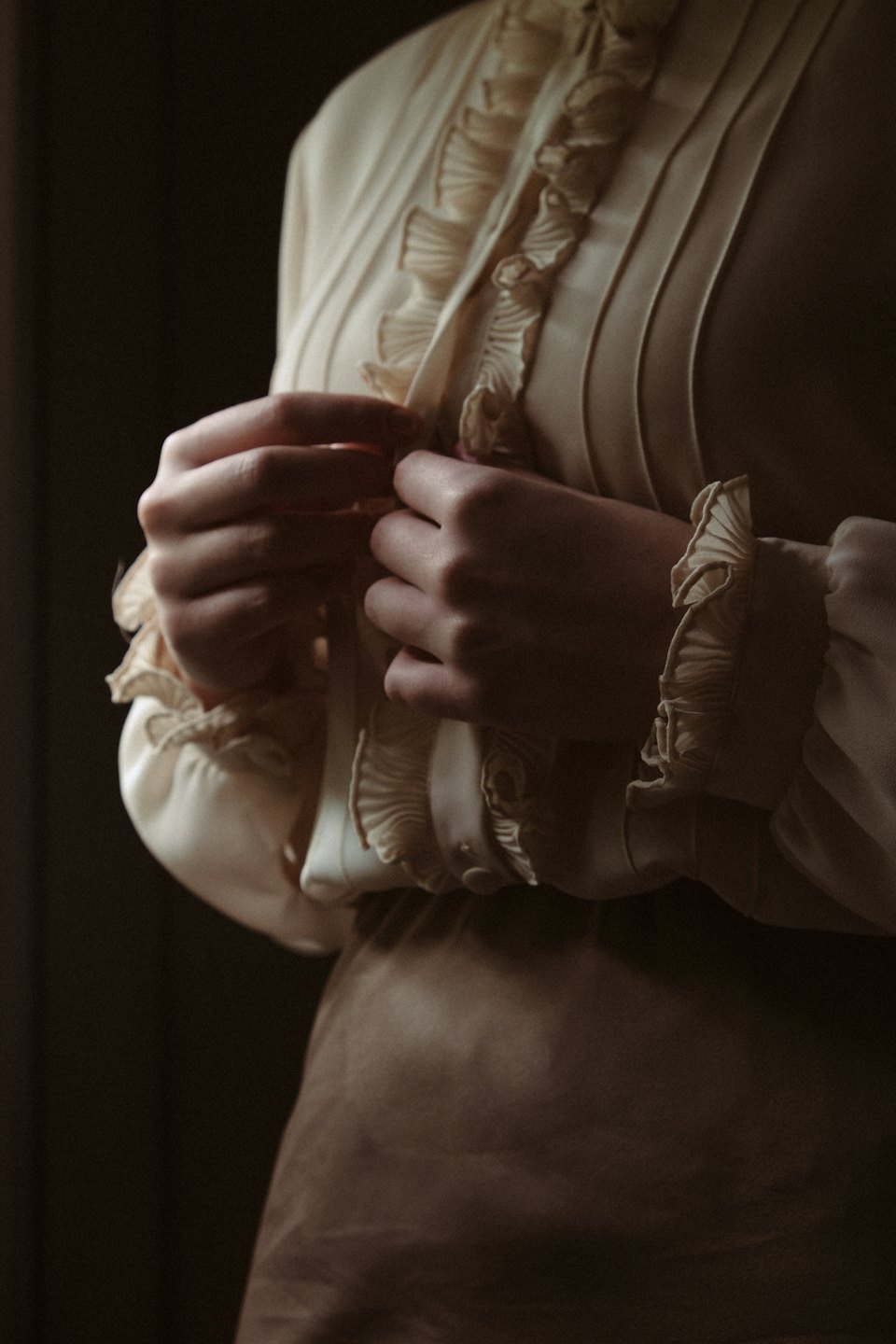 Hands buttoning the front of a cream-colored ruffled blouse.